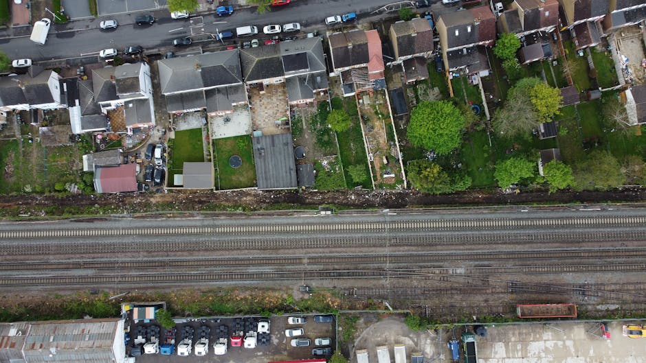 An aerial view of a residential area showing several garden plots and backyards separated by fences, with lush green trees and flowering plants. The gardens feature trimmed lawns, flower beds, and pav